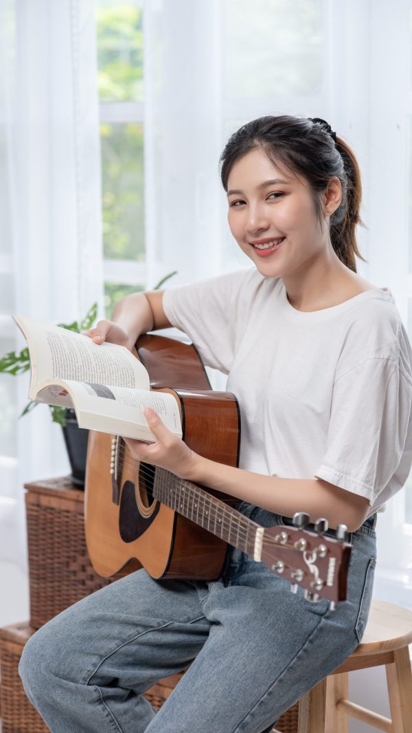 The girl is sitting and playing the guitar on the chair.